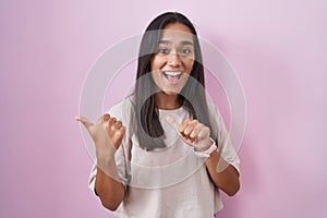 Young hispanic woman standing over pink background pointing to the back behind with hand and thumbs up, smiling confident