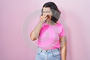 Young hispanic woman standing over pink background smelling something stinky and disgusting, intolerable smell, holding breath