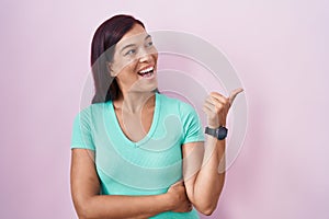 Young hispanic woman standing over pink background smiling with happy face looking and pointing to the side with thumb up