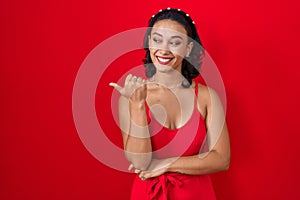 Young hispanic woman standing over red background smiling with happy face looking and pointing to the side with thumb up