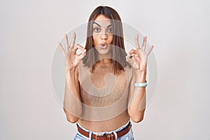 Young hispanic woman standing over white background looking surprised and shocked doing ok approval symbol with fingers