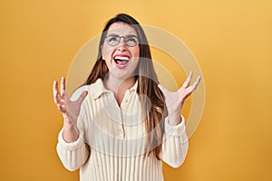 Young hispanic woman standing over yellow background crazy and mad shouting and yelling with aggressive expression and arms raised