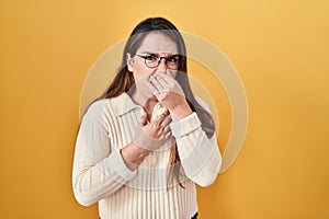 Young hispanic woman standing over yellow background smelling something stinky and disgusting, intolerable smell, holding breath