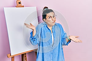 Young hispanic woman standing by painter easel stand clueless and confused expression with arms and hands raised