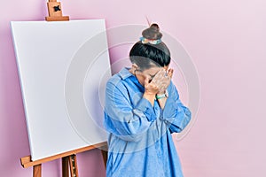 Young hispanic woman standing by painter easel stand with sad expression covering face with hands while crying