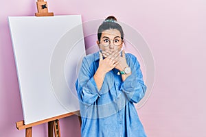 Young hispanic woman standing by painter easel stand shocked covering mouth with hands for mistake