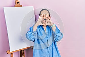 Young hispanic woman standing by painter easel stand shouting angry out loud with hands over mouth