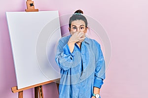 Young hispanic woman standing by painter easel stand smelling something stinky and disgusting, intolerable smell, holding breath