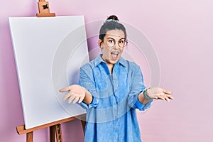 Young hispanic woman standing by painter easel stand smiling cheerful offering hands giving assistance and acceptance