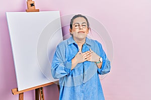 Young hispanic woman standing by painter easel stand smiling with hands on chest with closed eyes and grateful gesture on face