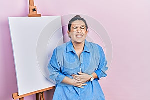 Young hispanic woman standing by painter easel stand smiling and laughing hard out loud because funny crazy joke with hands on