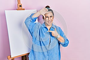 Young hispanic woman standing by painter easel stand smiling making frame with hands and fingers with happy face