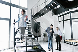 Young hispanic woman standing on the stairs and office colleagues busy working on computer in modern office in Mexico