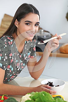Young hispanic woman or student cooking in kitchen. Girl tasting fresh salad while sitting at the table