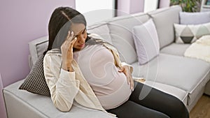 Young hispanic woman suffering a serious headache during pregnancy, sitting at home and massaging belly stressfully