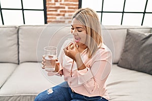 Young hispanic woman taking pill sitting on sofa at home