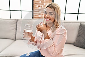 Young hispanic woman taking pill sitting on sofa at home