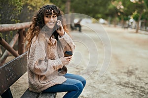 Young hispanic woman talking on the smartphone and drinking coffee at the park