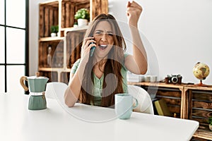 Young hispanic woman talking on the smartphone sitting on the table at home annoyed and frustrated shouting with anger, yelling