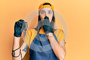 Young hispanic woman tattoo artist wearing professional uniform and gloves holding tattooer machine covering mouth with hand,