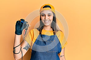 Young hispanic woman tattoo artist wearing professional uniform and gloves holding tattooer machine looking positive and happy