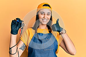 Young hispanic woman tattoo artist wearing professional uniform and gloves holding tattooer machine smiling happy and positive,