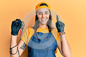 Young hispanic woman tattoo artist wearing professional uniform and gloves holding tattooer machine smiling with an idea or