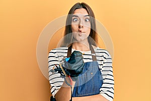 Young hispanic woman tattoo artist wearing professional uniform and gloves making fish face with mouth and squinting eyes, crazy