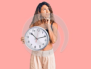 Young hispanic woman with tattoo holding big clock serious face thinking about question with hand on chin, thoughtful about