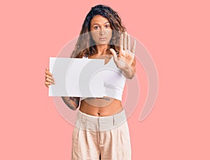 Young hispanic woman with tattoo holding blank empty paper with open hand doing stop sign with serious and confident expression,