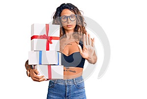 Young hispanic woman with tattoo holding gifts with open hand doing stop sign with serious and confident expression, defense
