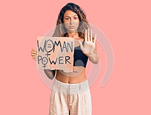 Young hispanic woman with tattoo holding woman power banner with open hand doing stop sign with serious and confident expression,