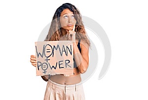 Young hispanic woman with tattoo holding woman power banner serious face thinking about question with hand on chin, thoughtful