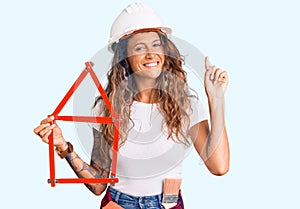 Young hispanic woman with tattoo wearing architect hardhat holding build project surprised with an idea or question pointing