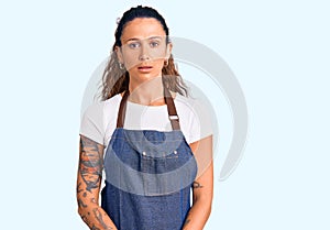 Young hispanic woman with tattoo wearing barber apron and gloves with serious expression on face