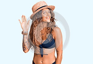 Young hispanic woman with tattoo wearing bikini and summer hat waiving saying hello happy and smiling, friendly welcome gesture