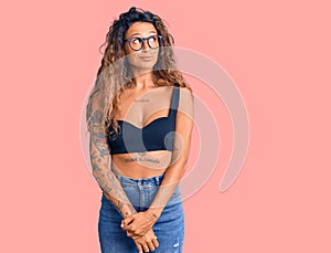 Young hispanic woman with tattoo wearing casual clothes and glasses smiling looking to the side and staring away thinking