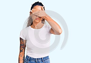 Young hispanic woman with tattoo wearing casual white tshirt covering eyes with hand, looking serious and sad