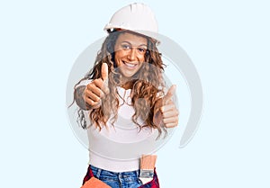 Young hispanic woman with tattoo wearing hardhat and builder clothes approving doing positive gesture with hand, thumbs up smiling