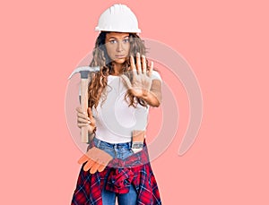 Young hispanic woman with tattoo wearing hardhat and builder clothes holding hammer with open hand doing stop sign with serious