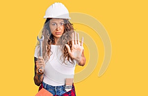 Young hispanic woman with tattoo wearing hardhat and builder clothes holding wrench with open hand doing stop sign with serious