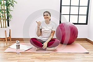 Young hispanic woman training at sport center smiling with happy face looking and pointing to the side with thumb up