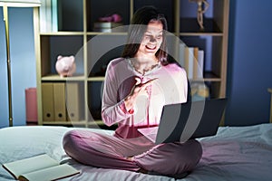 Young hispanic woman using computer laptop on the bed cheerful with a smile on face pointing with hand and finger up to the side