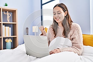 Young hispanic woman using computer laptop on the bed pointing thumb up to the side smiling happy with open mouth