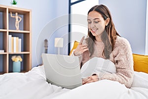 Young hispanic woman using computer laptop on the bed smiling happy pointing with hand and finger