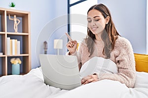 Young hispanic woman using computer laptop on the bed smiling happy pointing with hand and finger to the side