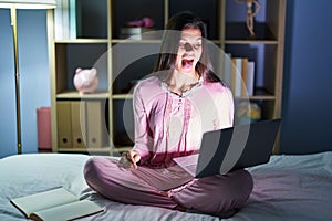 Young hispanic woman using computer laptop on the bed sticking tongue out happy with funny expression