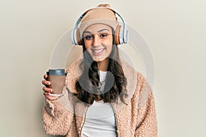 Young hispanic woman using headphones drinking coffee looking positive and happy standing and smiling with a confident smile