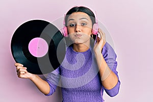 Young hispanic woman using headphones holding vinyl disc puffing cheeks with funny face