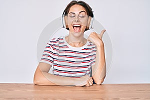 Young hispanic woman using headphones sitting on the table smiling with happy face looking and pointing to the side with thumb up